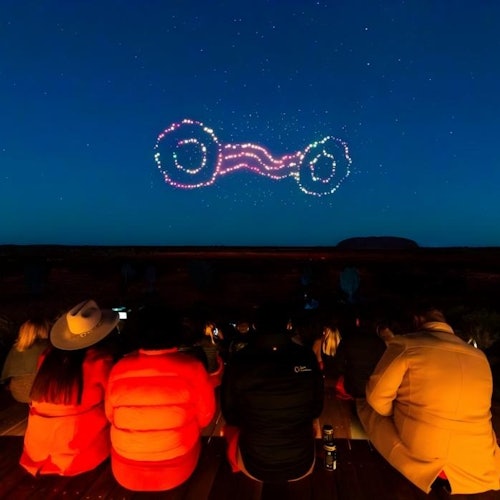 A group of people watch a drone light show forming interconnected rings in the night sky.