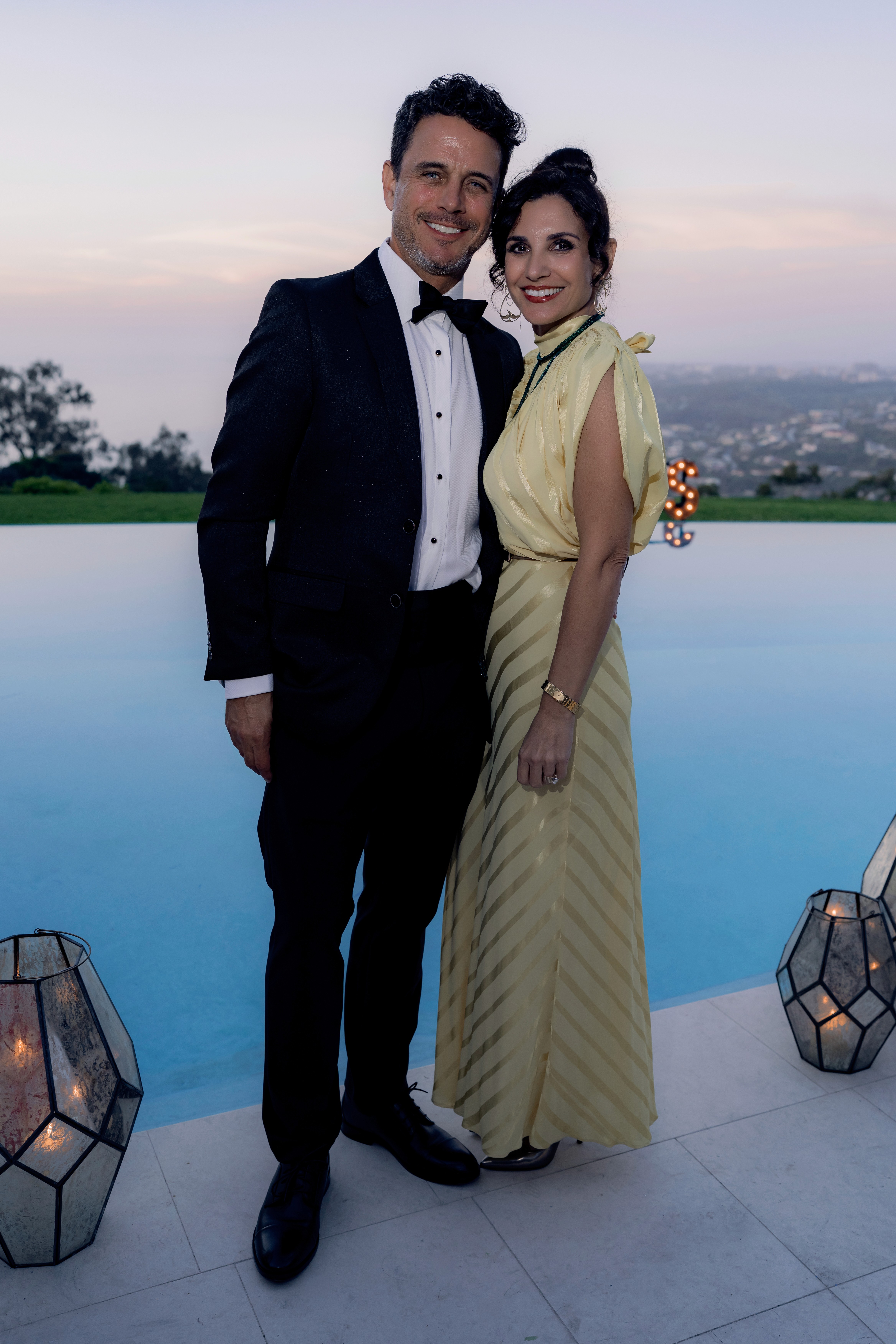 A couple posing together at an outdoor evening event, the man in a black tuxedo and the woman in a pale yellow gown.