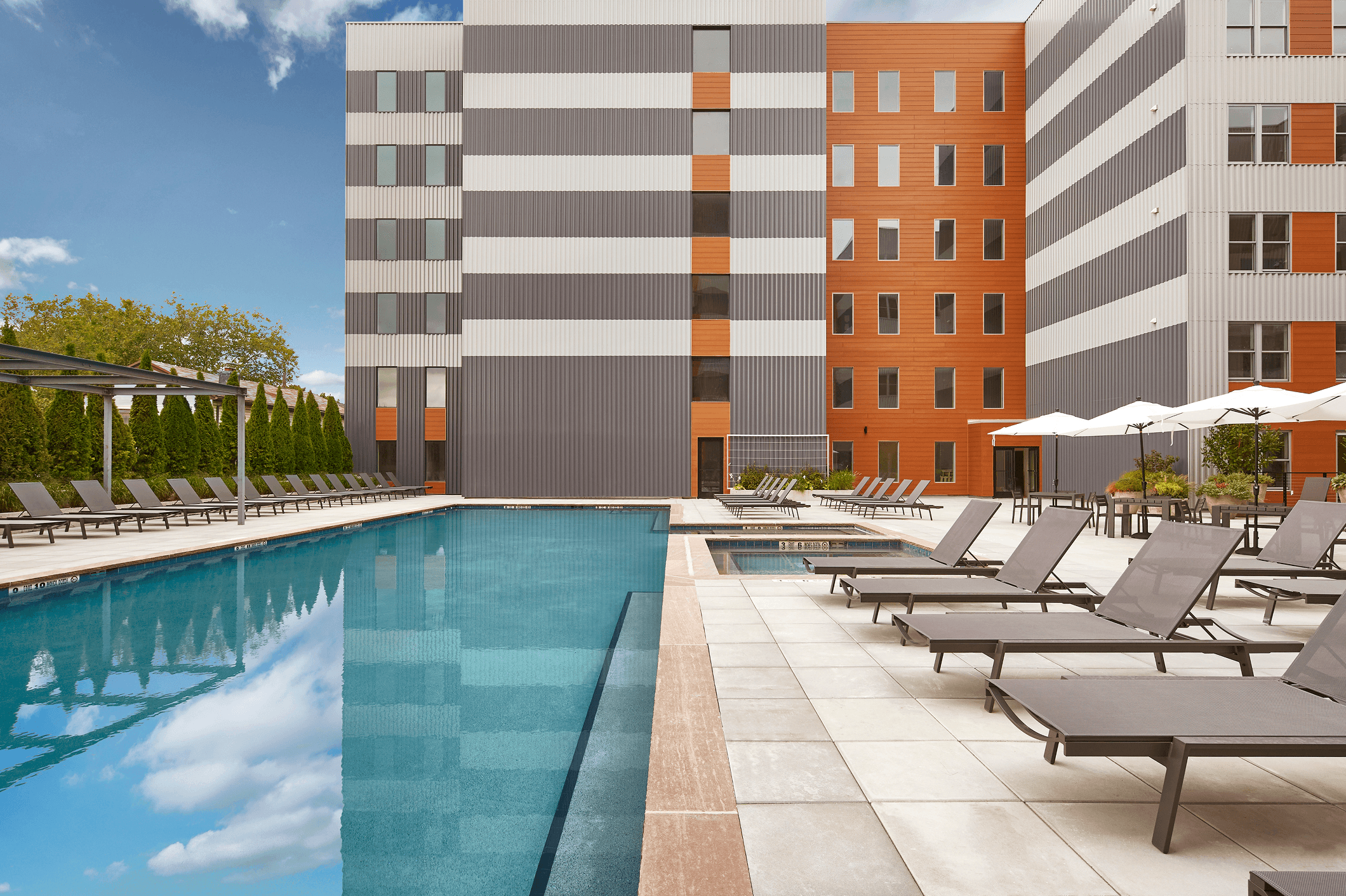 Modern apartment complex with a swimming pool and lounge chairs under a clear blue sky.