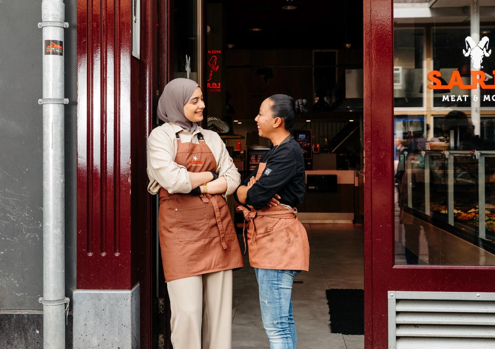 A bright, modern dining room featuring a long light wood table set with several matching wooden chairs and white cushions, illuminated by natural light streaming through large windows overlooking a lush green and sunlit yard.