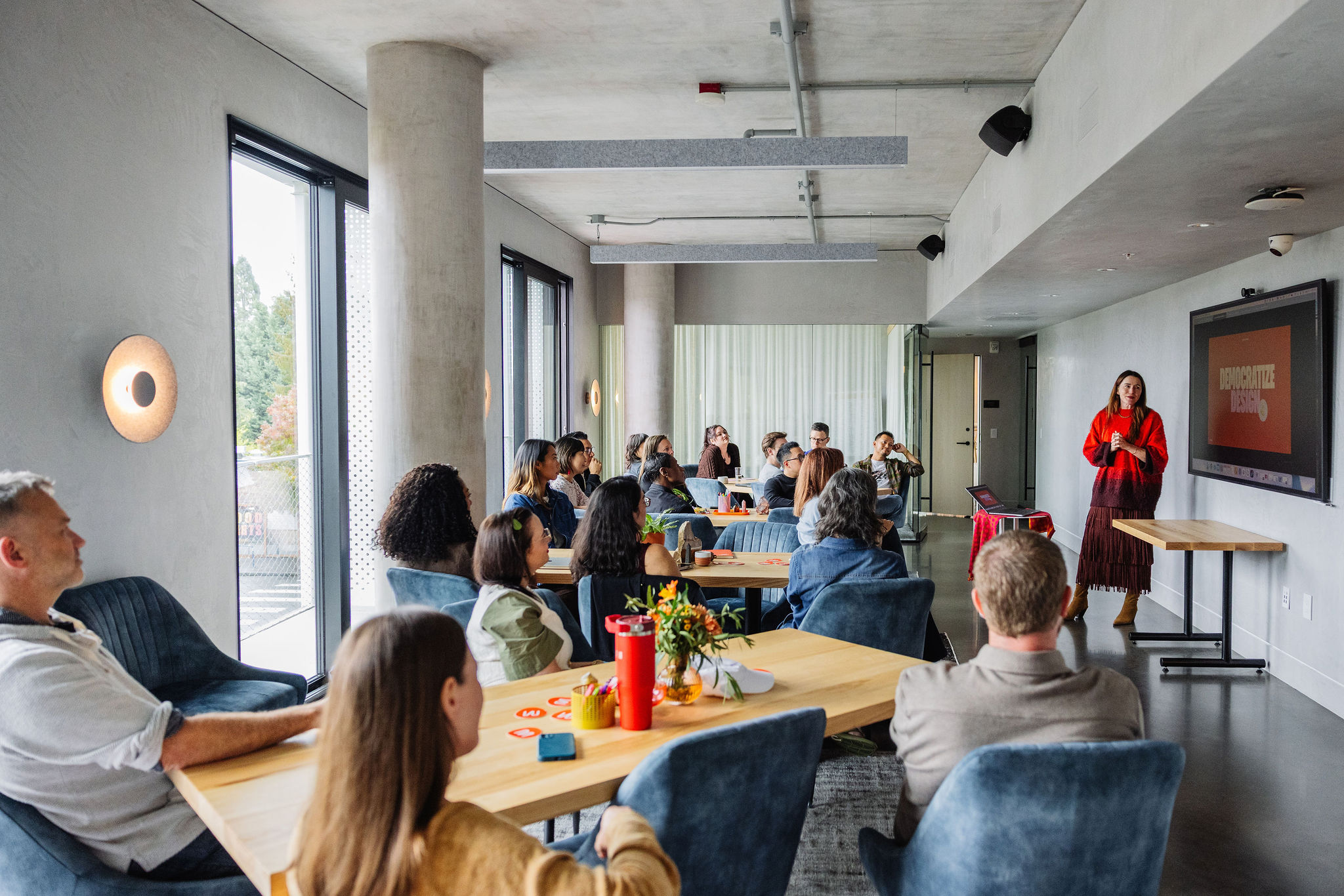 Wide view of a group meeting in a modern office, with attendees seated at tables and a speaker presenting at the front of the room.