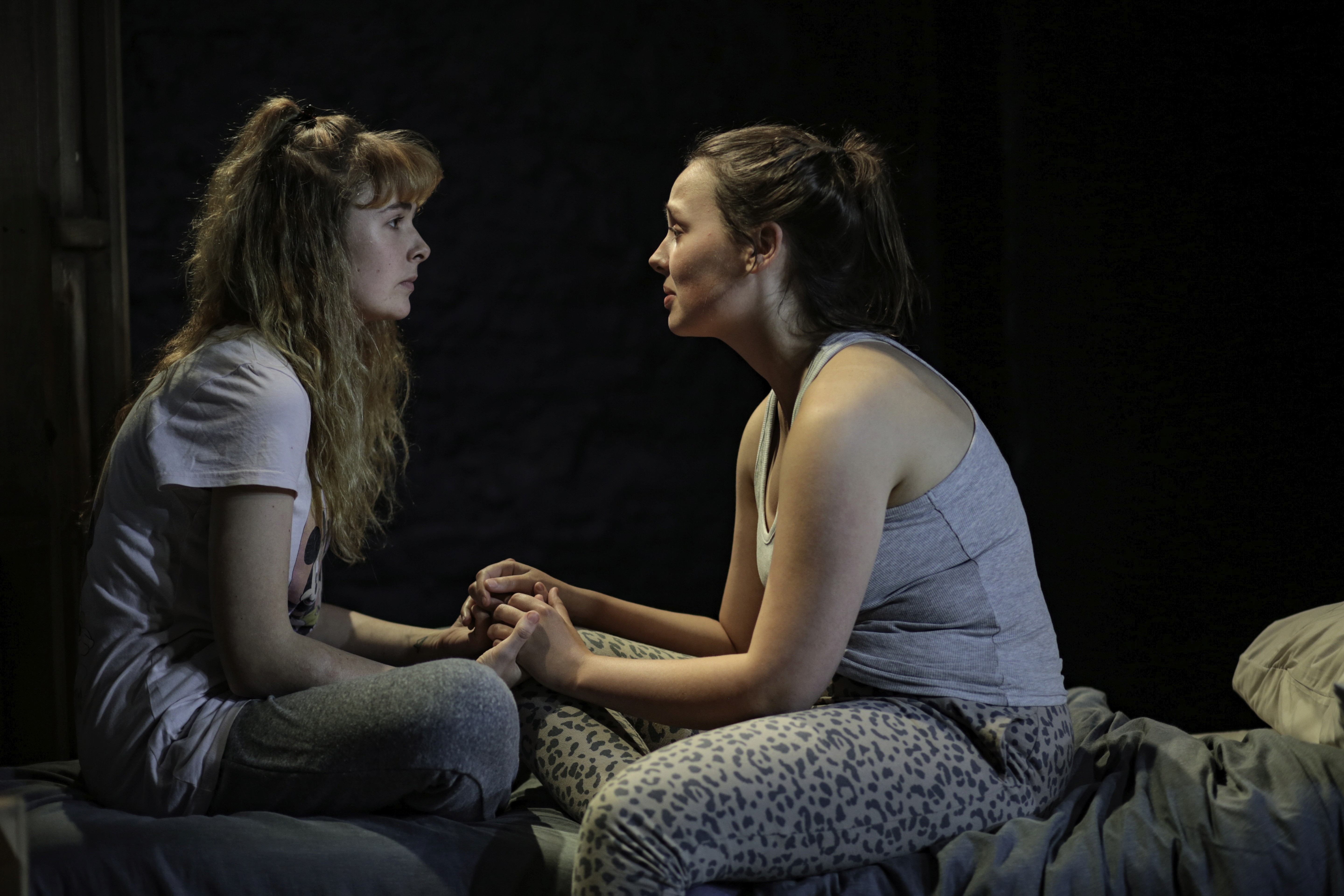 Two young women sit closely on a bed in pyjamas, holding hands and making eye contact. They share a quiet, intimate moment that feels vulnerable and supportive against the dark set.