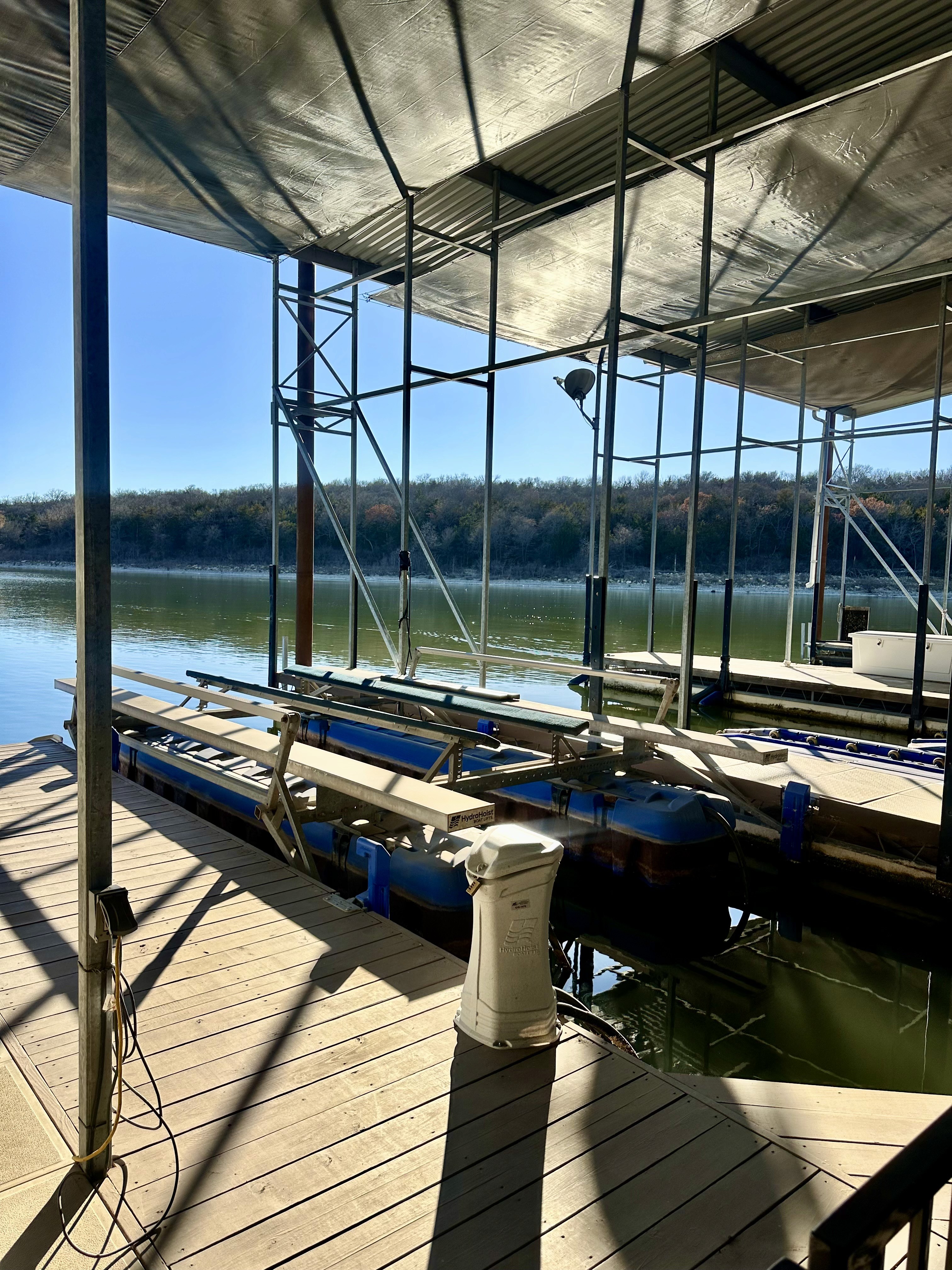 A sunlit marina dock extends over a tranquil river, surrounded by green foliage, with several boats moored beneath a metal canopy.