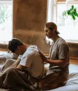Man met pet legt zijn handen op de rug van een man die met een deken op een matras zit