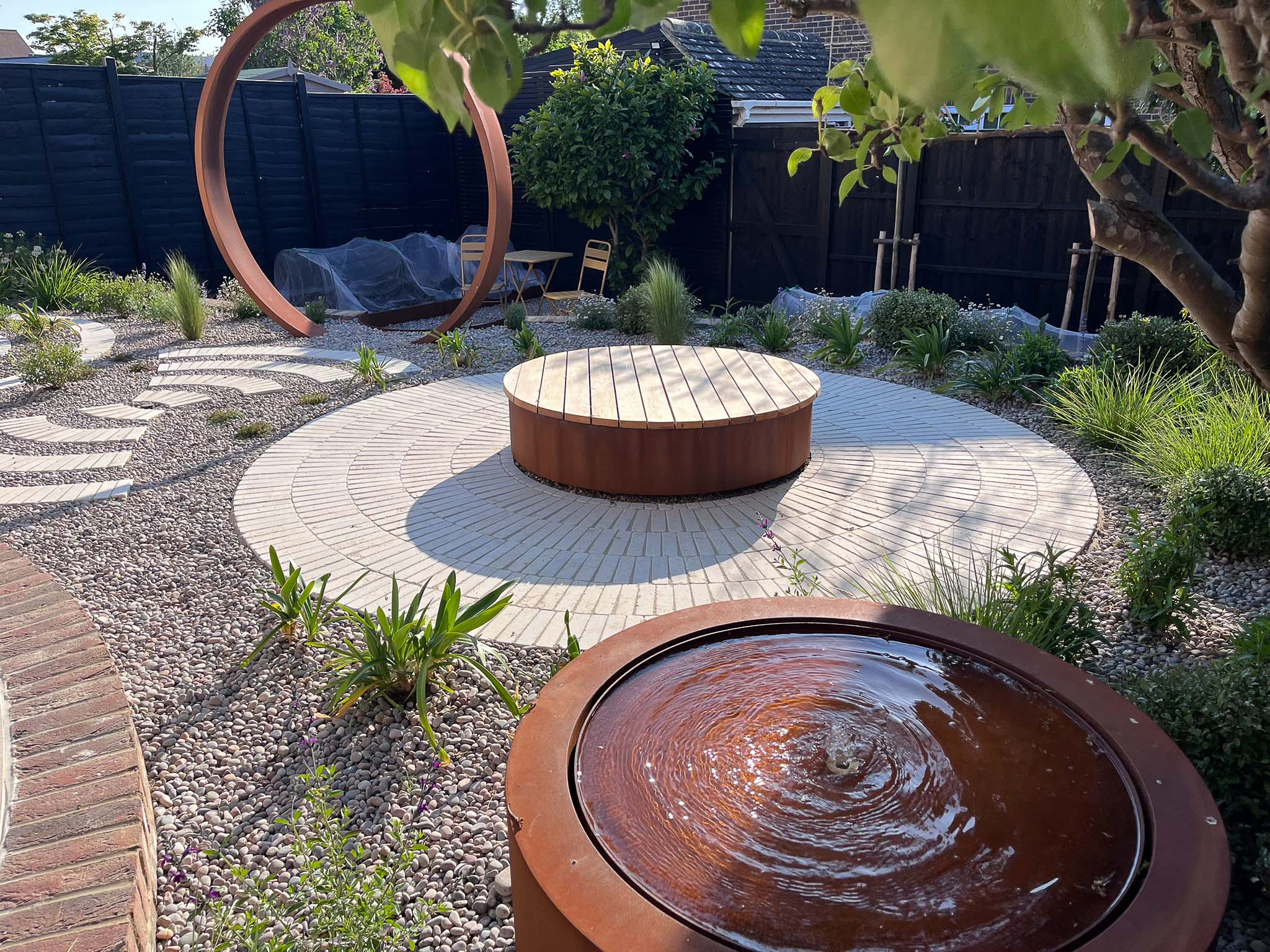 A landscaped garden featuring circular stone seating, a metal ring sculpture, and surrounding greenery.