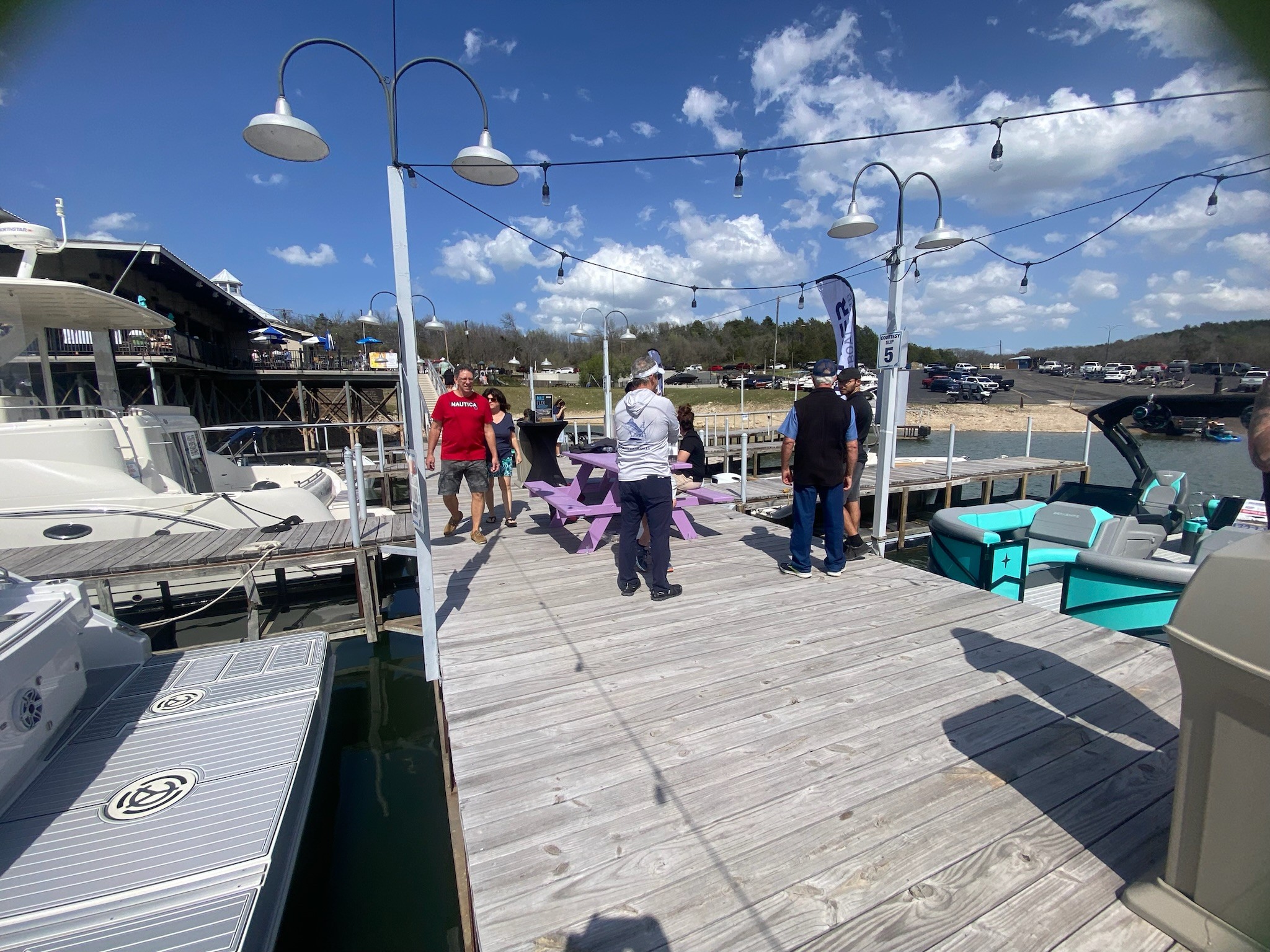 A sunny day at a marina features people walking on a wooden dock lined with boats, benches, and light poles under a partly cloudy sky.