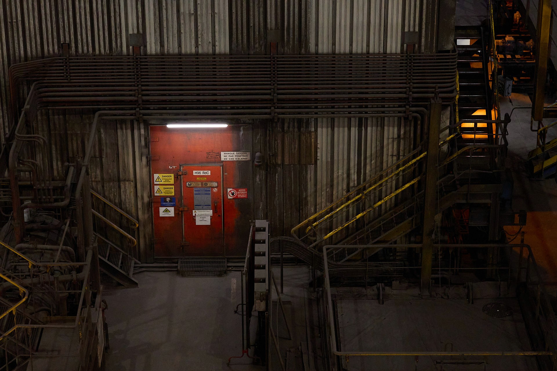 Reportage industriel sur une coulée continue : vue en plongée d'une porte de sécurité rouge sale, éclairée par un néon et entourée d'un réseau complexe de tuyauterie et de panneaux de danger dans une usine.