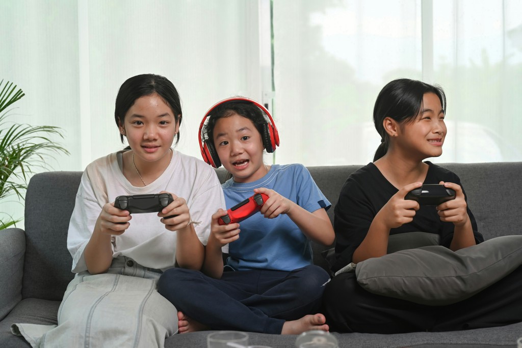 Three children enthusiastically playing video games on a couch, with one child wearing red headphones and holding a controller, while the other two are also engrossed in the game.