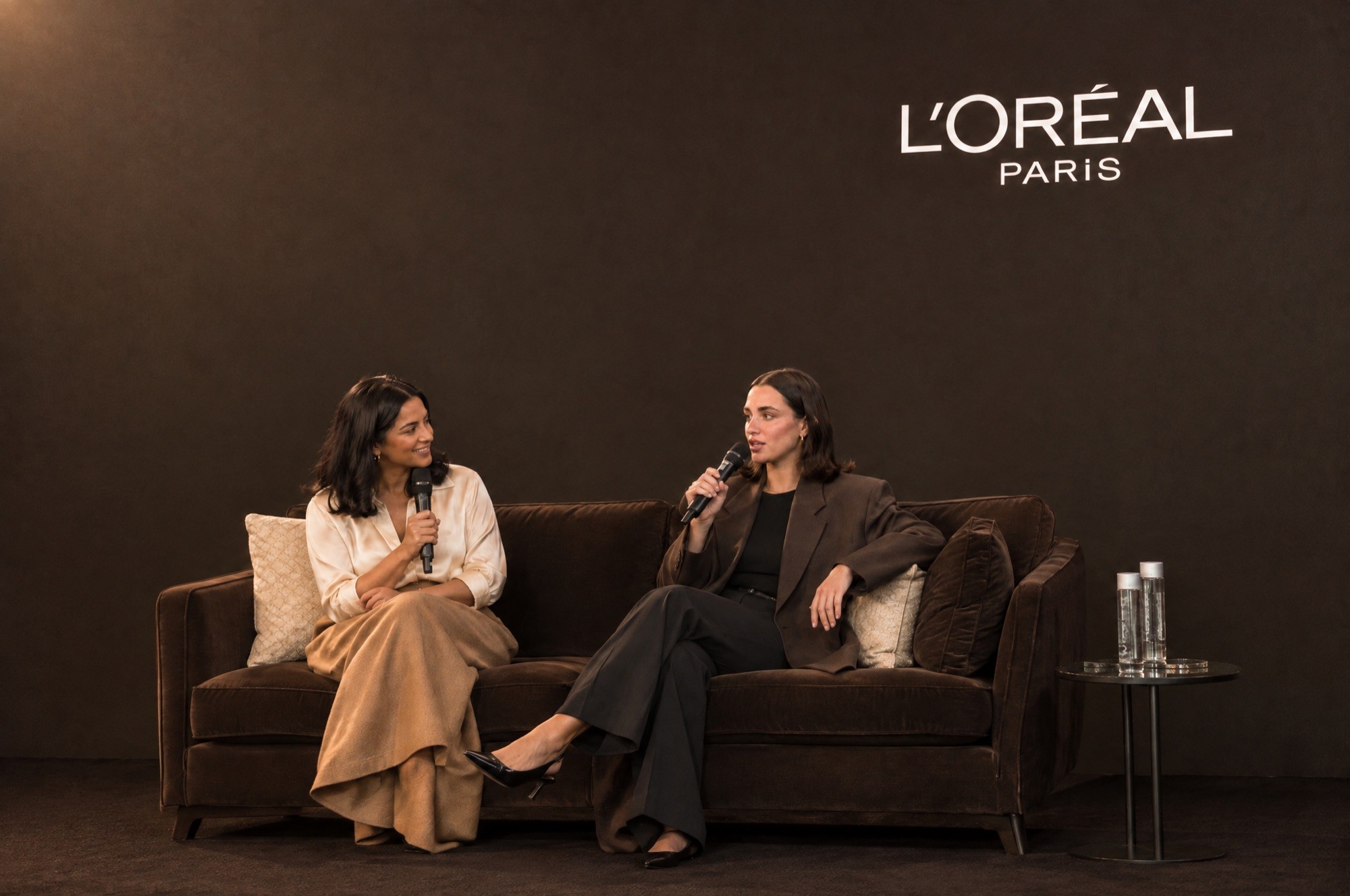 Margaux Ellis of SEEN speaking on a panel at a L'Oréal Paris press event, seated on a sofa beside the host.