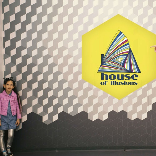 Дете в розово яке стои пред стената с шарка, на която има шестоъгълен знак "Къща на илюзиите".