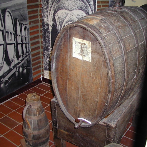 An old wooden barrel with a spigot sits on a stand next to a smaller wooden keg, with historic barrel photos in the background.
