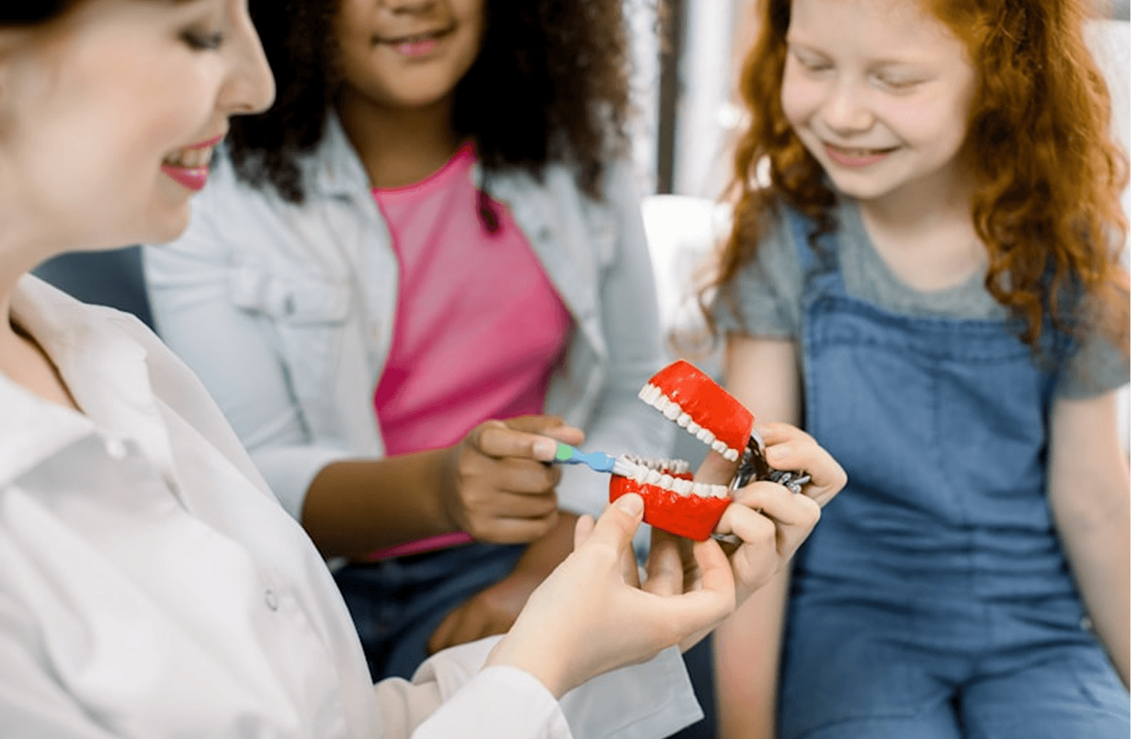 A dental professional demonstrates proper tooth brushing technique on a red and white dental model to two smiling children, emphasizing the importance of oral hygiene in a bright, friendly environment.