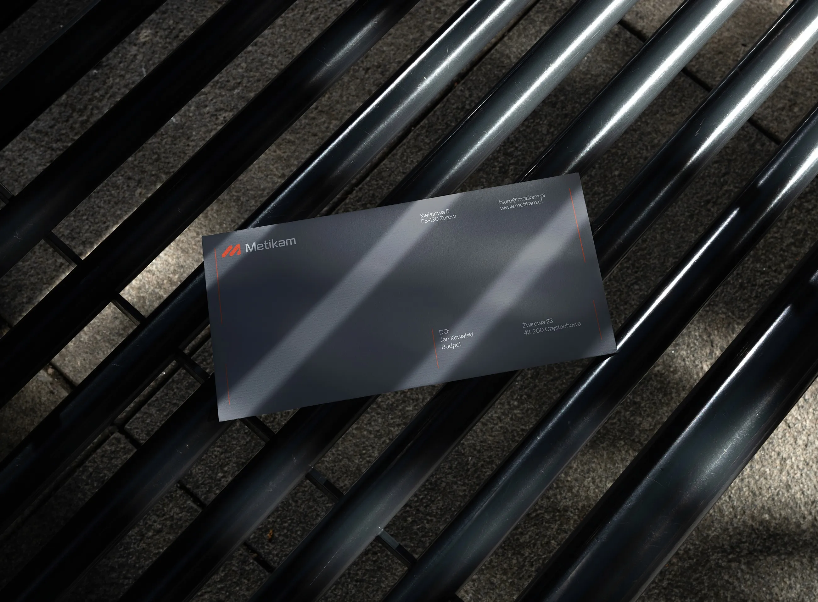 Dark grey Metikam DL envelope with orange logo on steel pipes, corporate stationery design