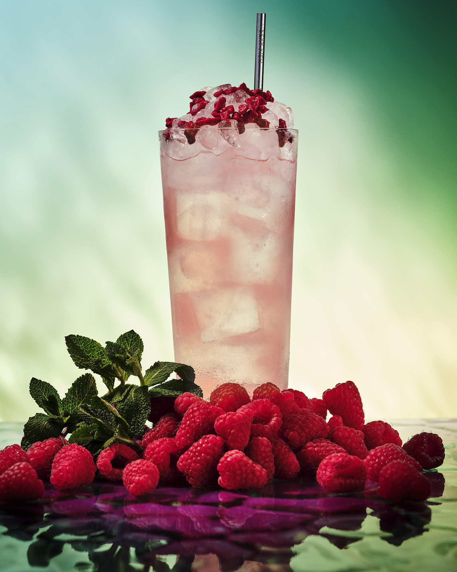 A tall glass of pink drink with ice, topped with raspberries, sits on a bed of fresh strawberries on a colorful background.