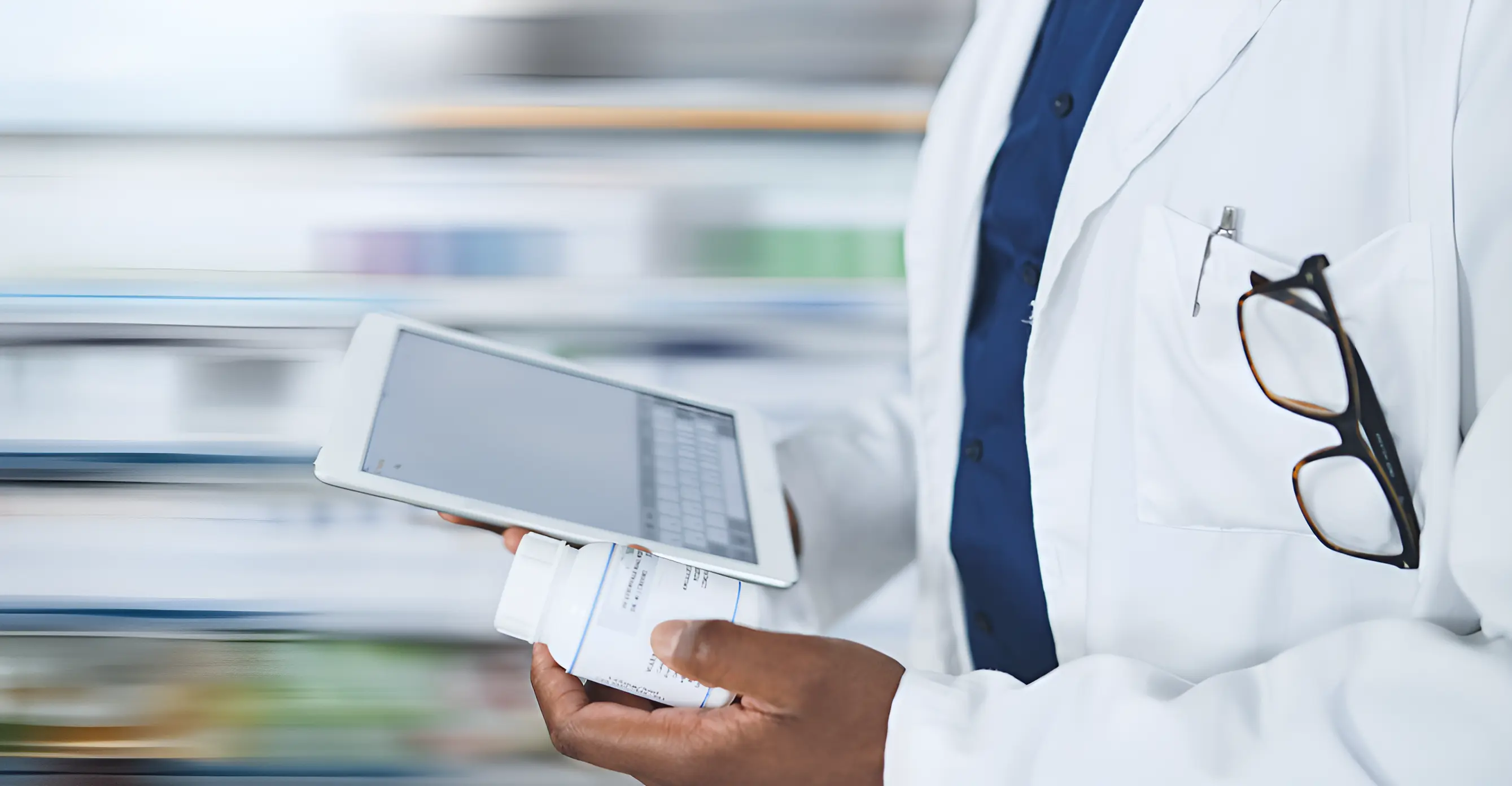 Pharmacist in a lab coat consulting a tablet while holding a medication bottle, with glasses in his pocket.