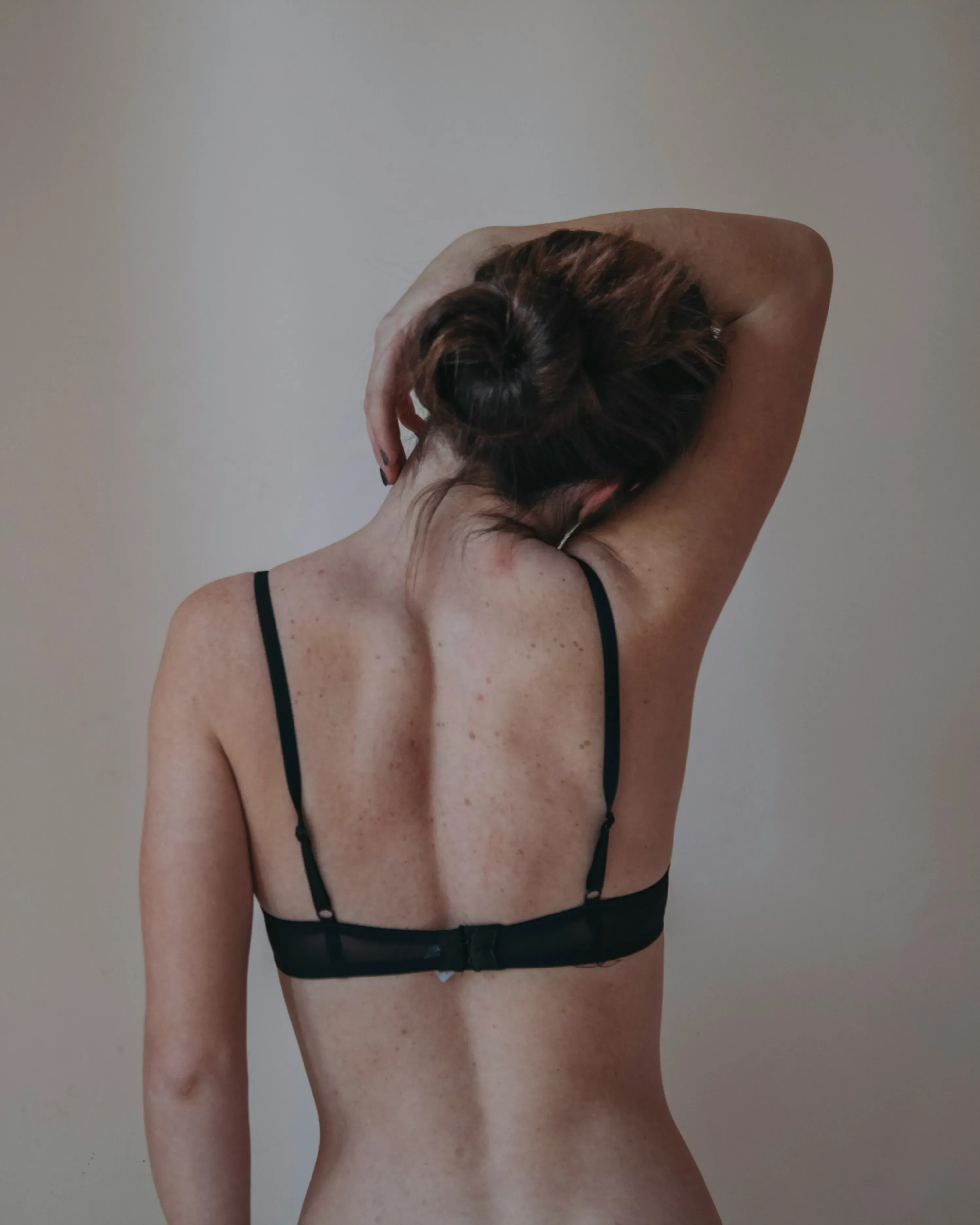 A person with short hair is seen from behind, wearing a black bra and holding their arm behind their head against a neutral wall.