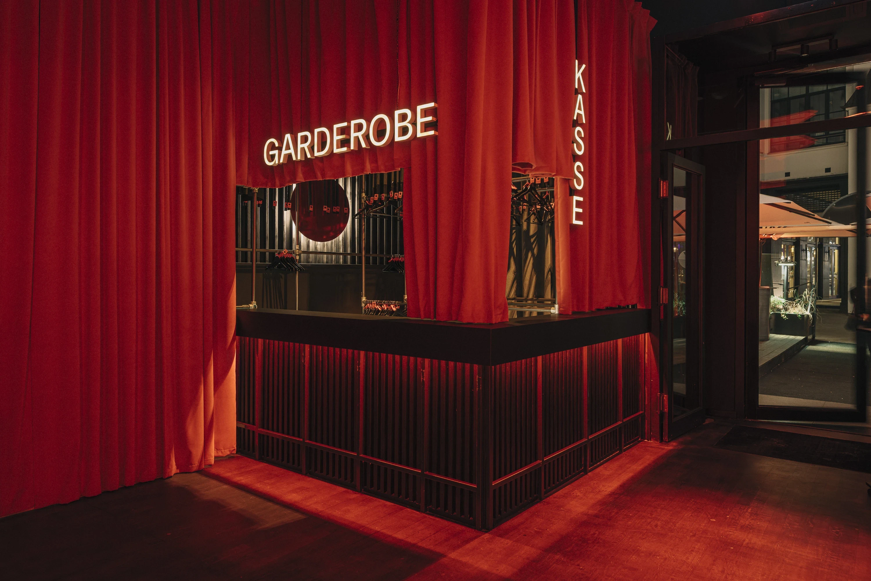 Entrance of NATE CLUB with red velvet curtains, a black slatted counter and illuminated Garderobe and Kasse lettering above the cloakroom.