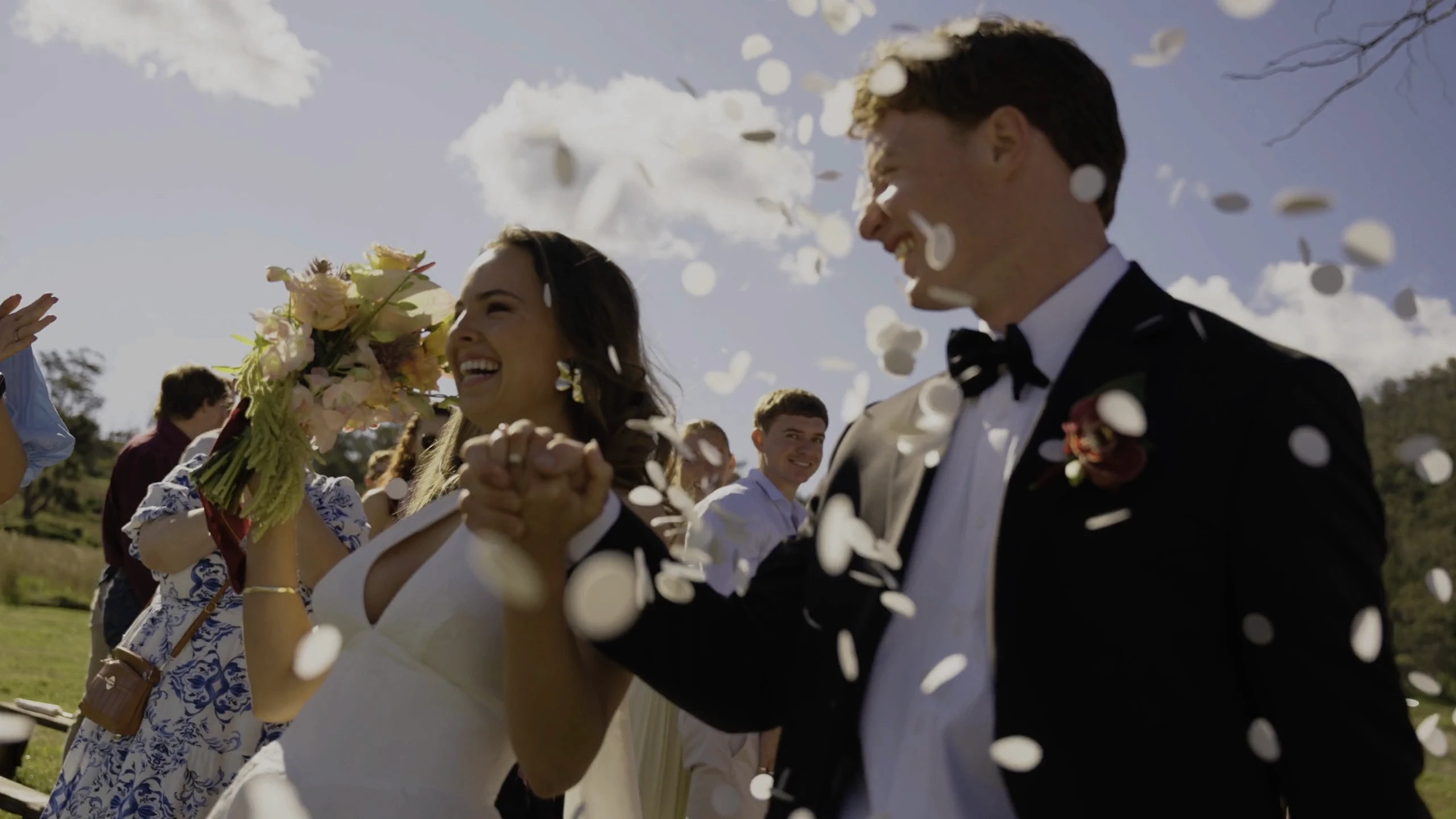A joyful bride and groom walk hand in hand under a shower of white petals, surrounded by well-wishers and a sunny, blue sky.