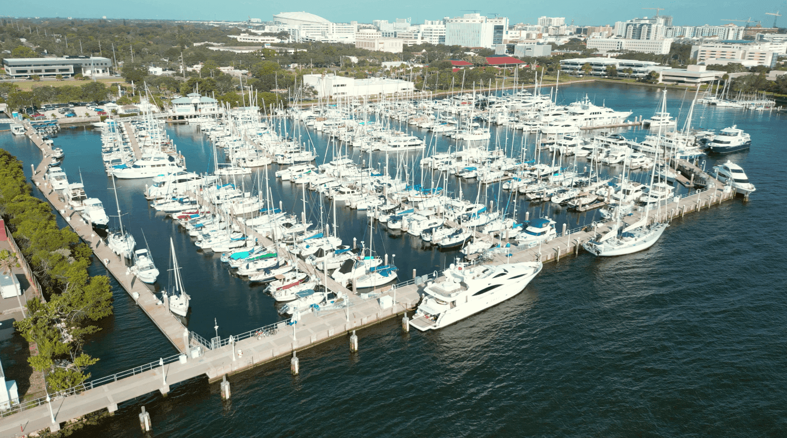 Arial view of a bustling marina filled with numerous sailboats and yachts, surrounded by a cityscape of white buildings and lush greenery, under a clear blue sky.