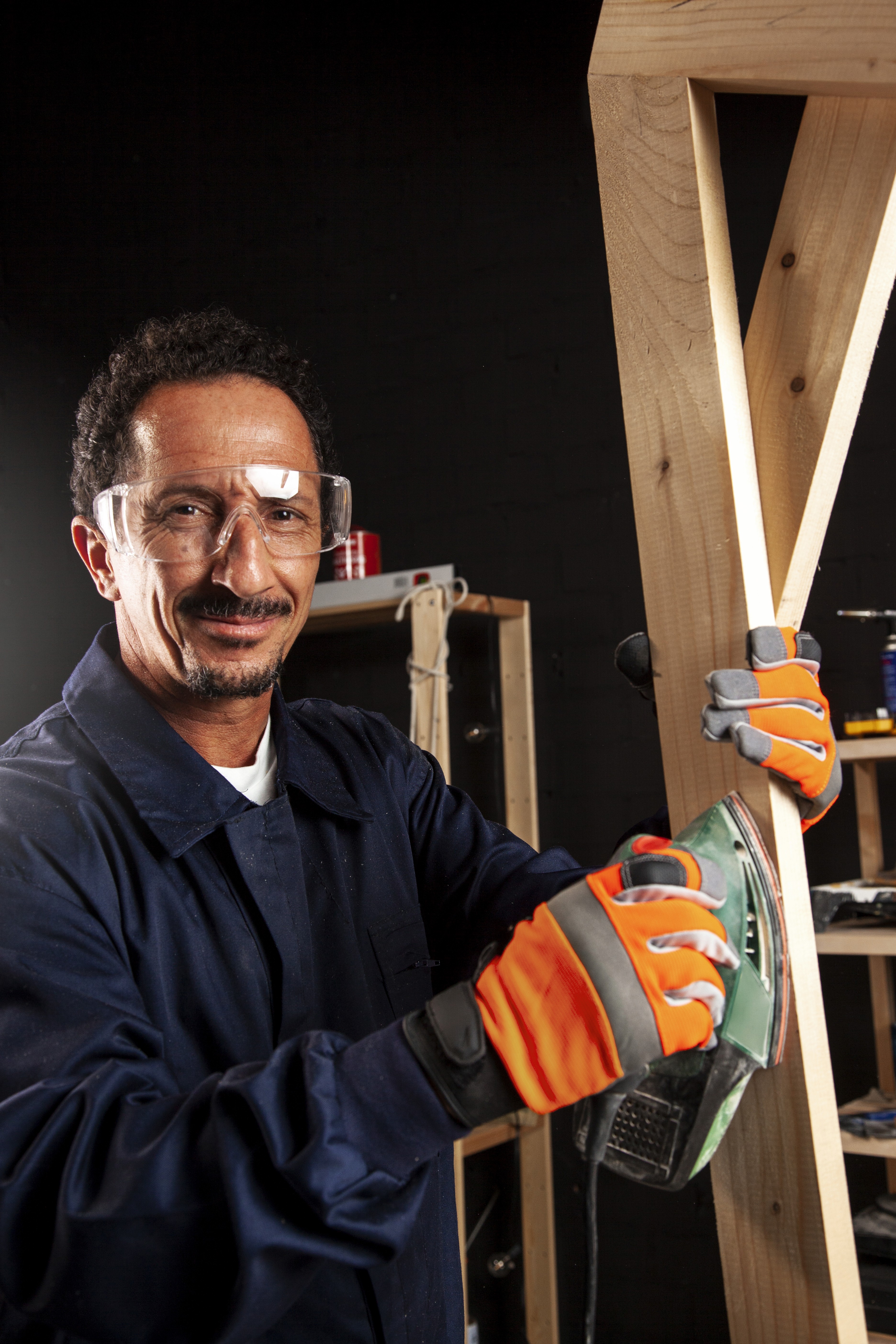A man sanding a wooden chair in a workshop, wearing gloves, a cap, and safety earmuffs.