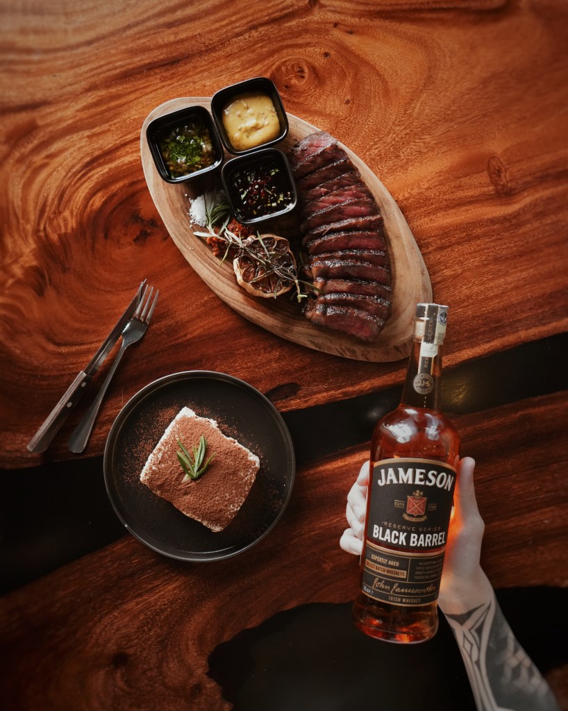 A wooden table displaying a platter of sliced steak accompanied by three small bowls of sauces, alongside a dessert plate of tiramisu topped with a sprig of rosemary, and a bottle of Jameson Black Barrel whiskey held by a hand with tattoos.