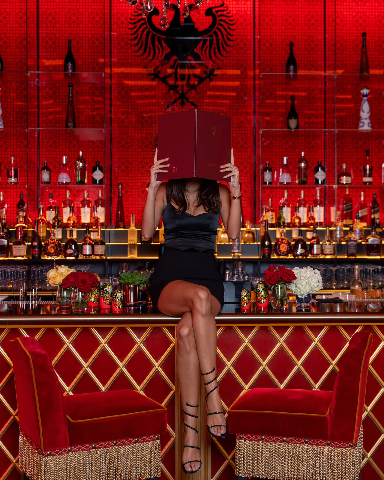 Woman in a black dress sitting on the red velvet bar at Raspoutine Dubai, hiding behind the menu, with the red mosaic back bar and double-headed eagle behind her