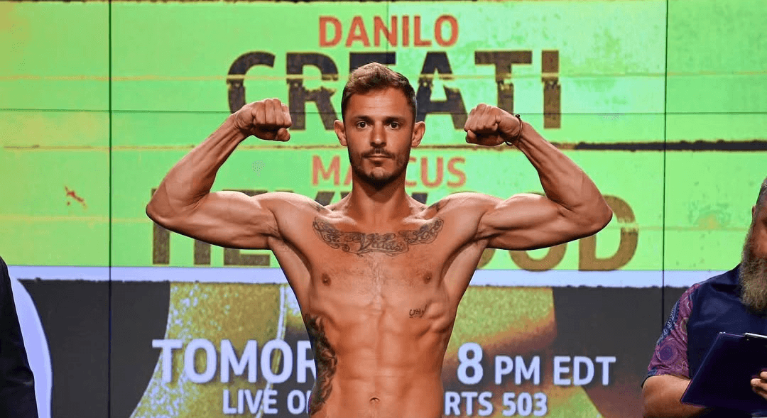Danilo Dash Creati flexing on stage during weigh-ins in front of a green screen background.