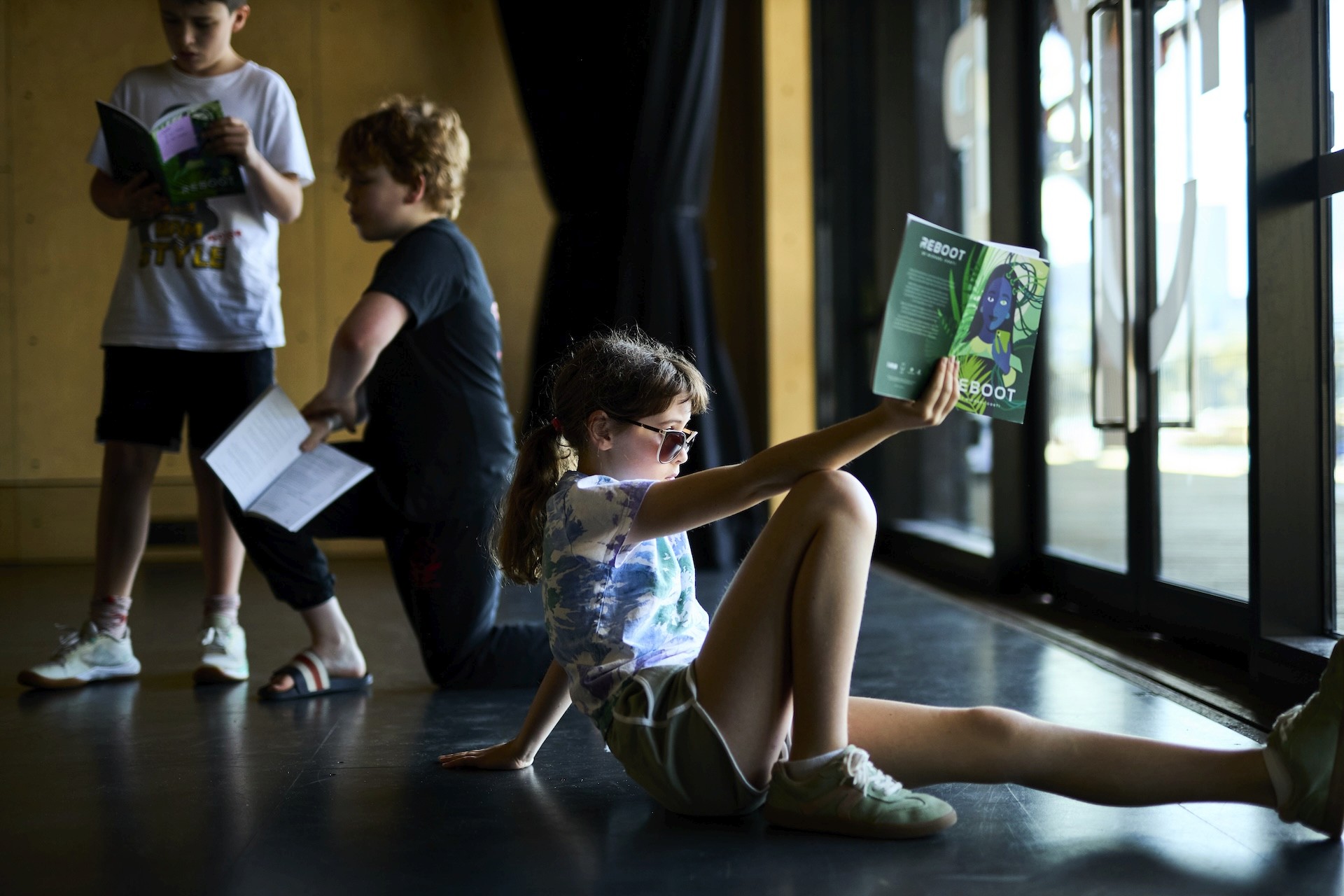 A child sits on the studio floor reading from a booklet while other children rehearse and read scripts nearby.