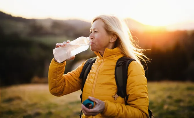 Active senior woman in a yellow jacket drinking water from a bottle while hiking outdoors at sunset.