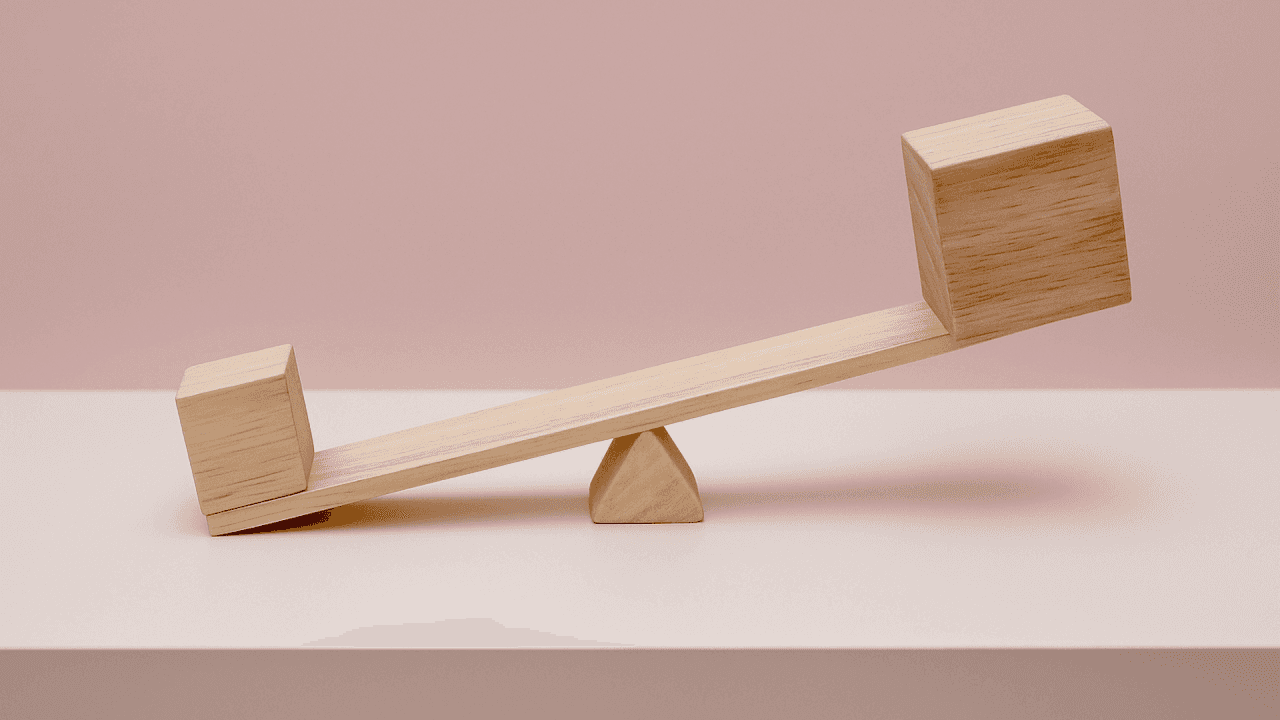 Wooden seesaw with a small block lifting a larger one, symbolizing leverage and efficiency, set against a soft pink background on a white surface.