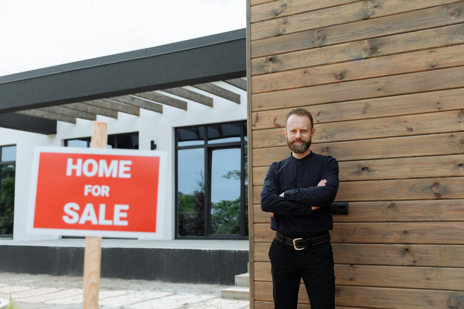 Bearded man in a black shirt with arms crossed beside a red Home For Sale sign outside a modern house
