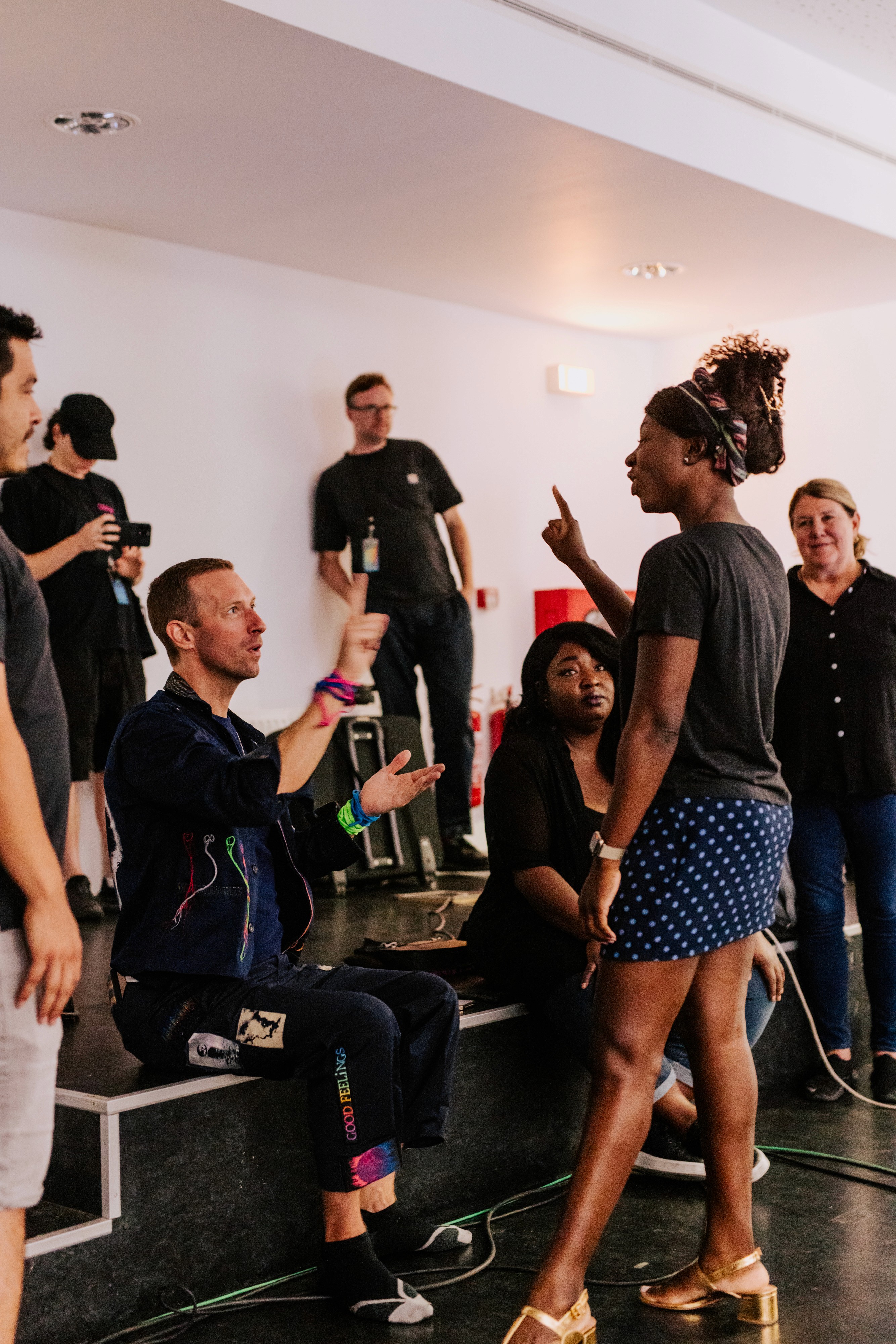 Natasha Ofili (a dark-skinned Black woman) teaching Chris Martin (a white, British man) a sign while others watch intently. 