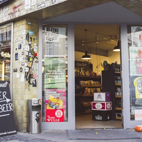 Pintu kaca terbuka di pintu masuk toko menampilkan rak-rak produk di dalam dan papan nama yang dipenuhi stiker di samping tempat sampah.
