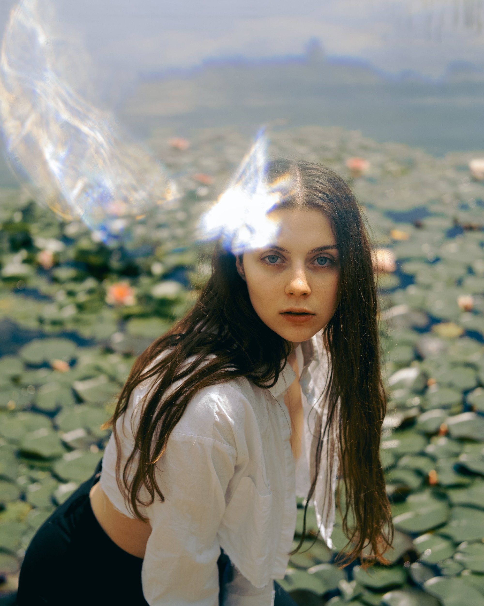 Person in white shirt near a pond with lily pads, looking intensely toward the viewer with light flare effects.