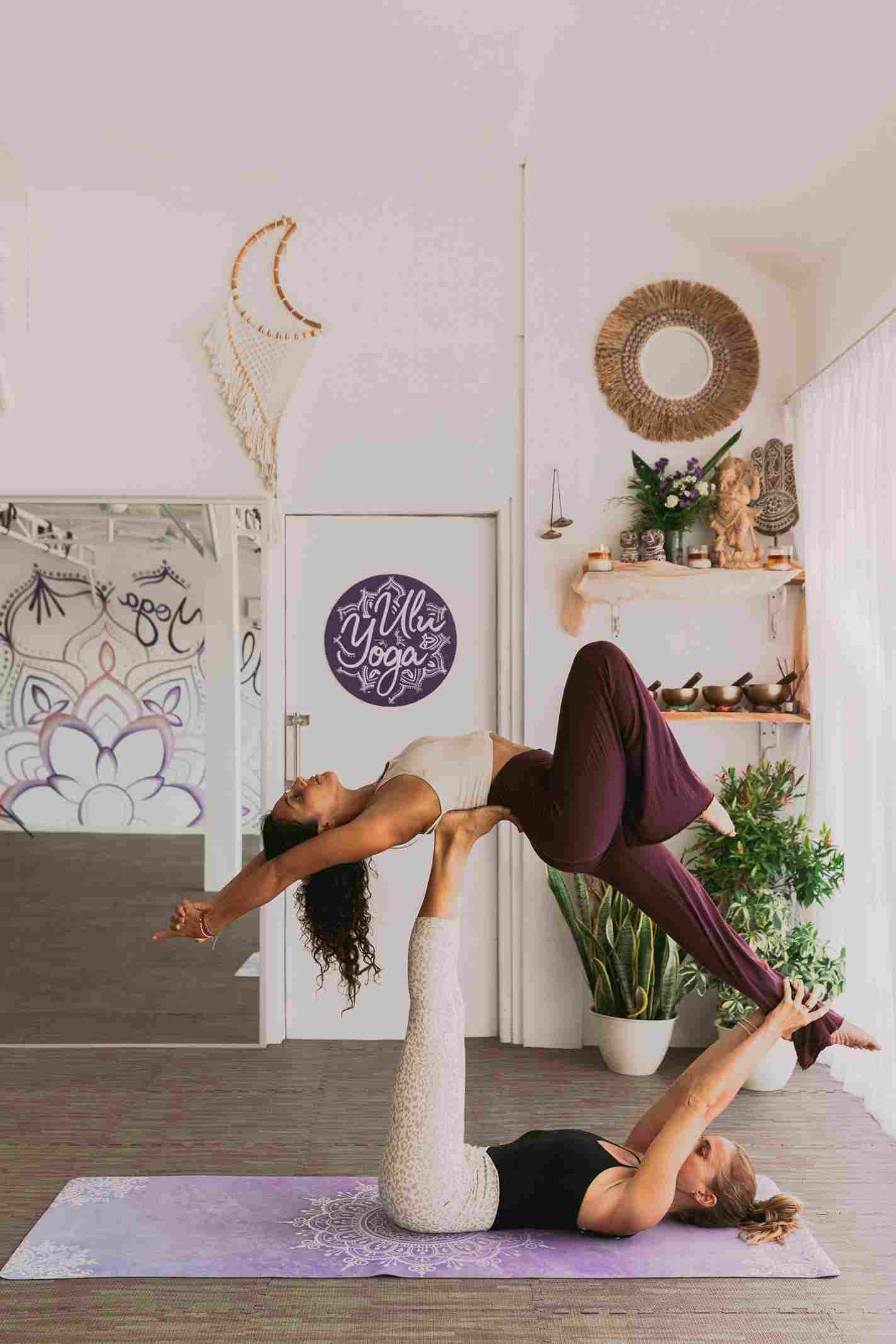 Two women practicing Acro yoga flying pose inside the Ulu Yoga studio with mandala wall art.