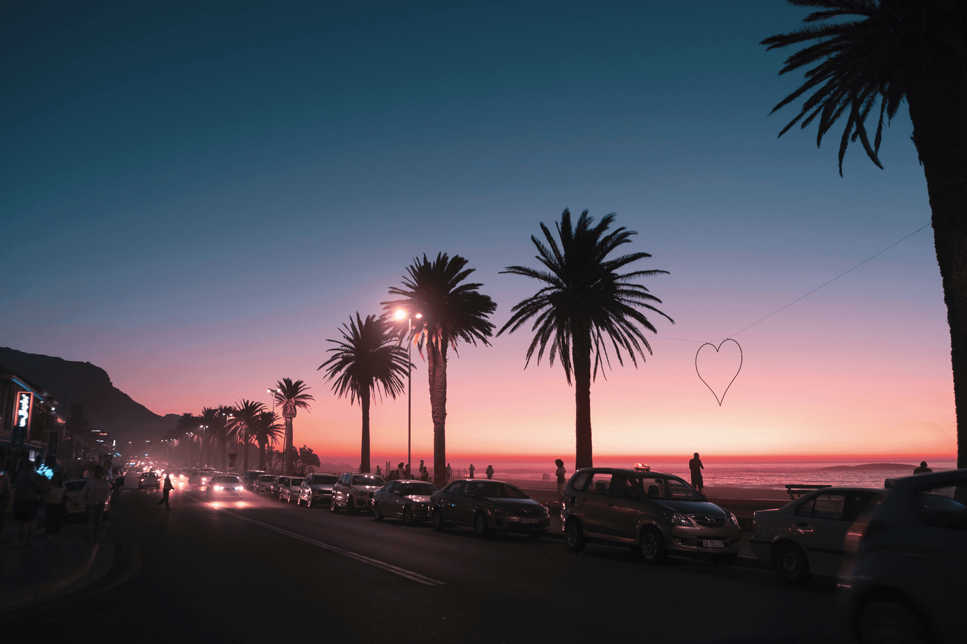 Sonnenuntergang an der Küste. Palmen säumen die Straße, Autos parken am Strand, Herz im Himmel.