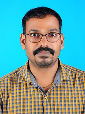 A woman with long wavy hair stands confidently in a bright office, wearing a white shirt and patterned blazer. The setting conveys professionalism.