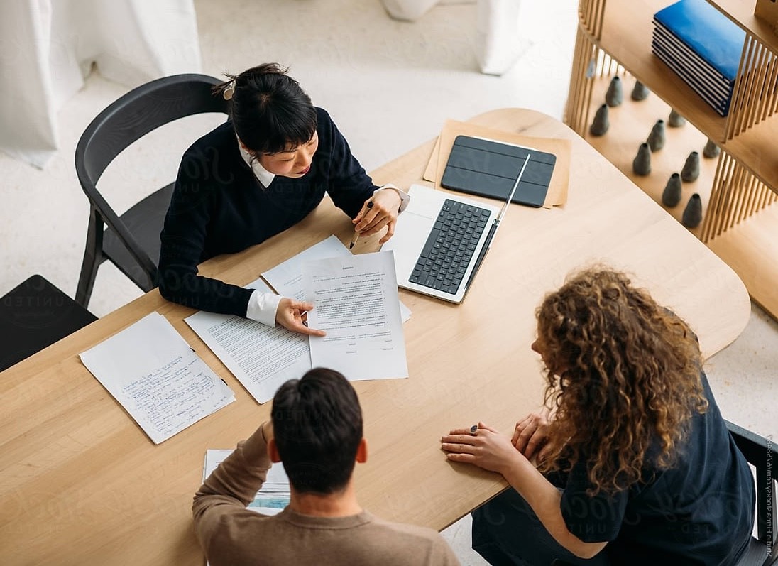 Young man in a green hoodie sitting at a table with a yellow phone in hand and a tablet in front of him, smiling at the camera.
