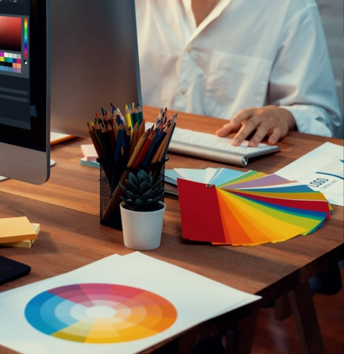 A person works at a desk with a computer, surrounded by colorful design printouts and art supplies.