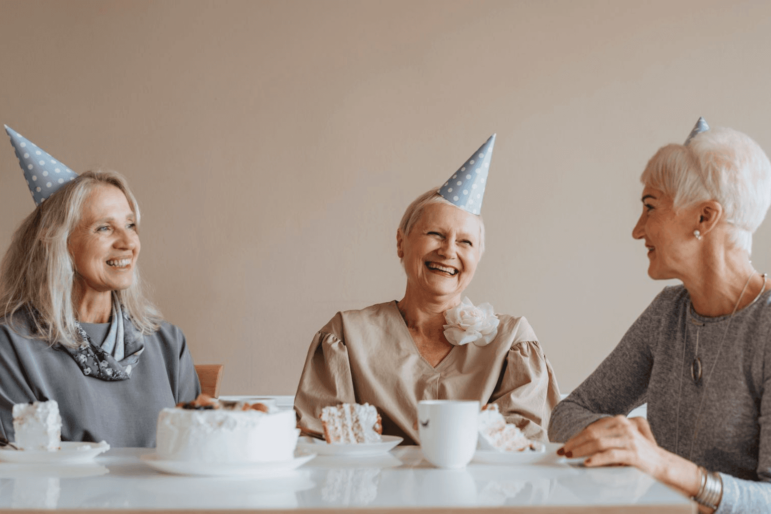 Three older women laughing over cake and coffee at a table together.