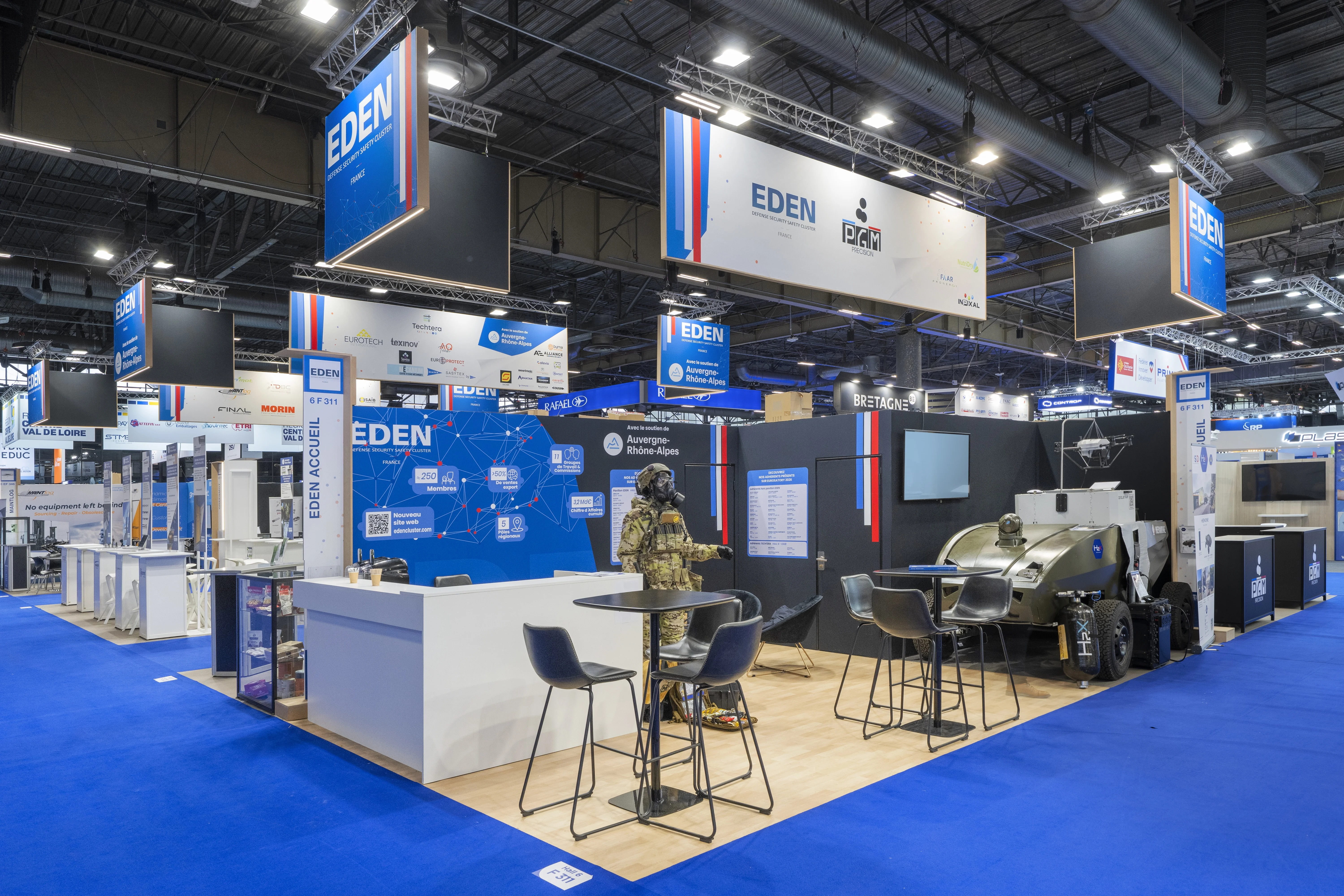 Stand EDEN Cluster conçu et réalisé par Expace à Eurosatory 2026 – pavillon collectif défense bleu-blanc-rouge regroupant les adhérents (SAIB, Sysnav, OBSAM, PGM…) avec vitrines produits, démonstrateurs et accueil central.