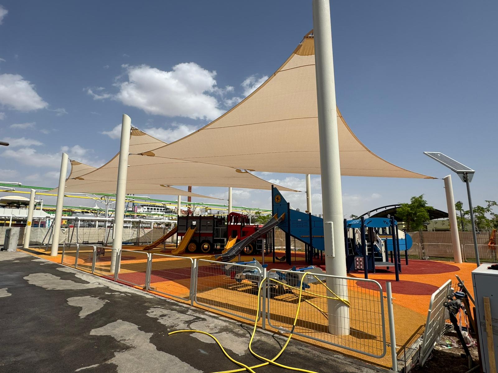 Playground with slides and climbing structure under a large triangular shade sail supported by metal poles.