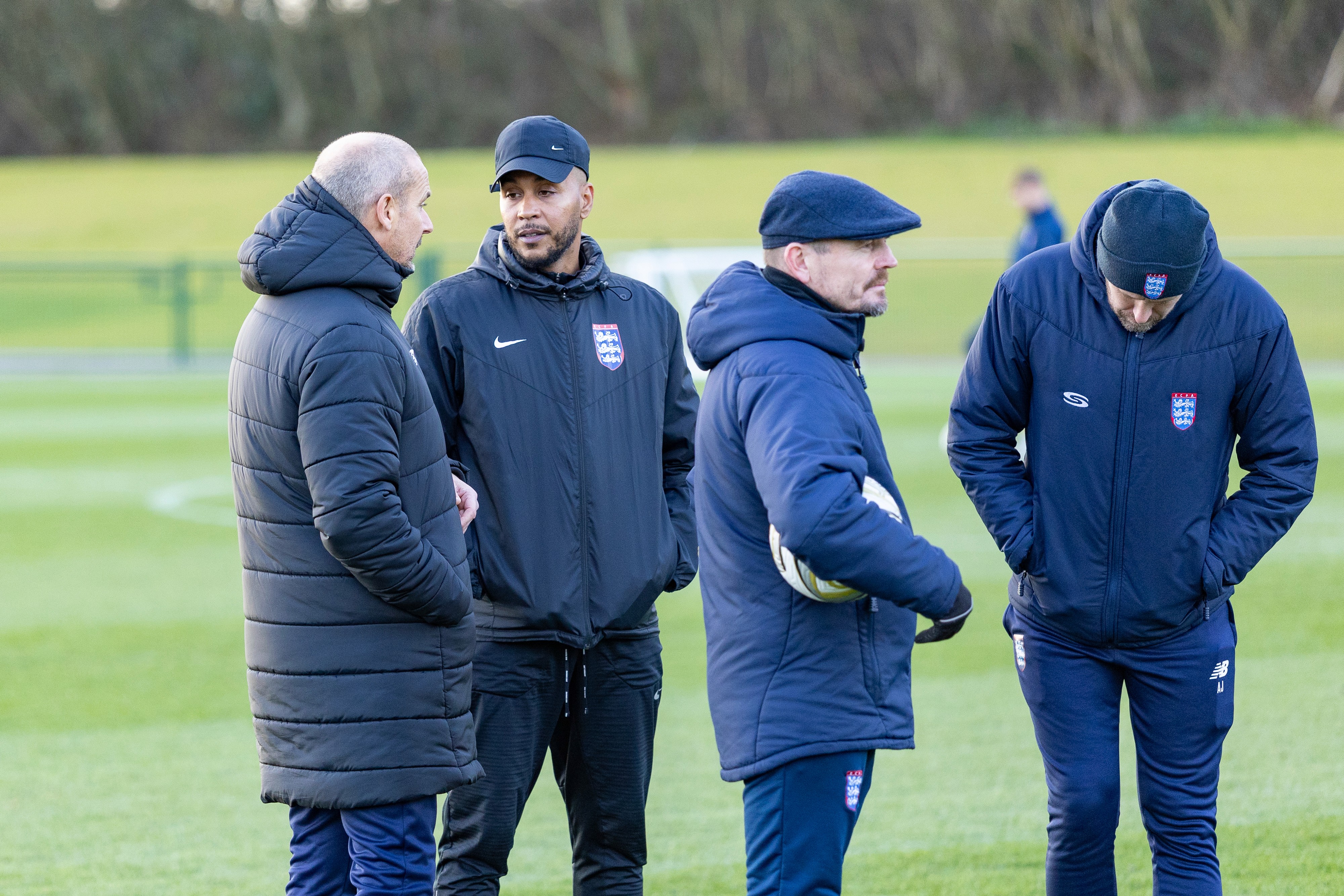Four individuals dressed in matching dark sports jackets and hats converse on a grassy field, with one holding a soccer ball, emphasizing a casual sports coaching or meeting scenario.