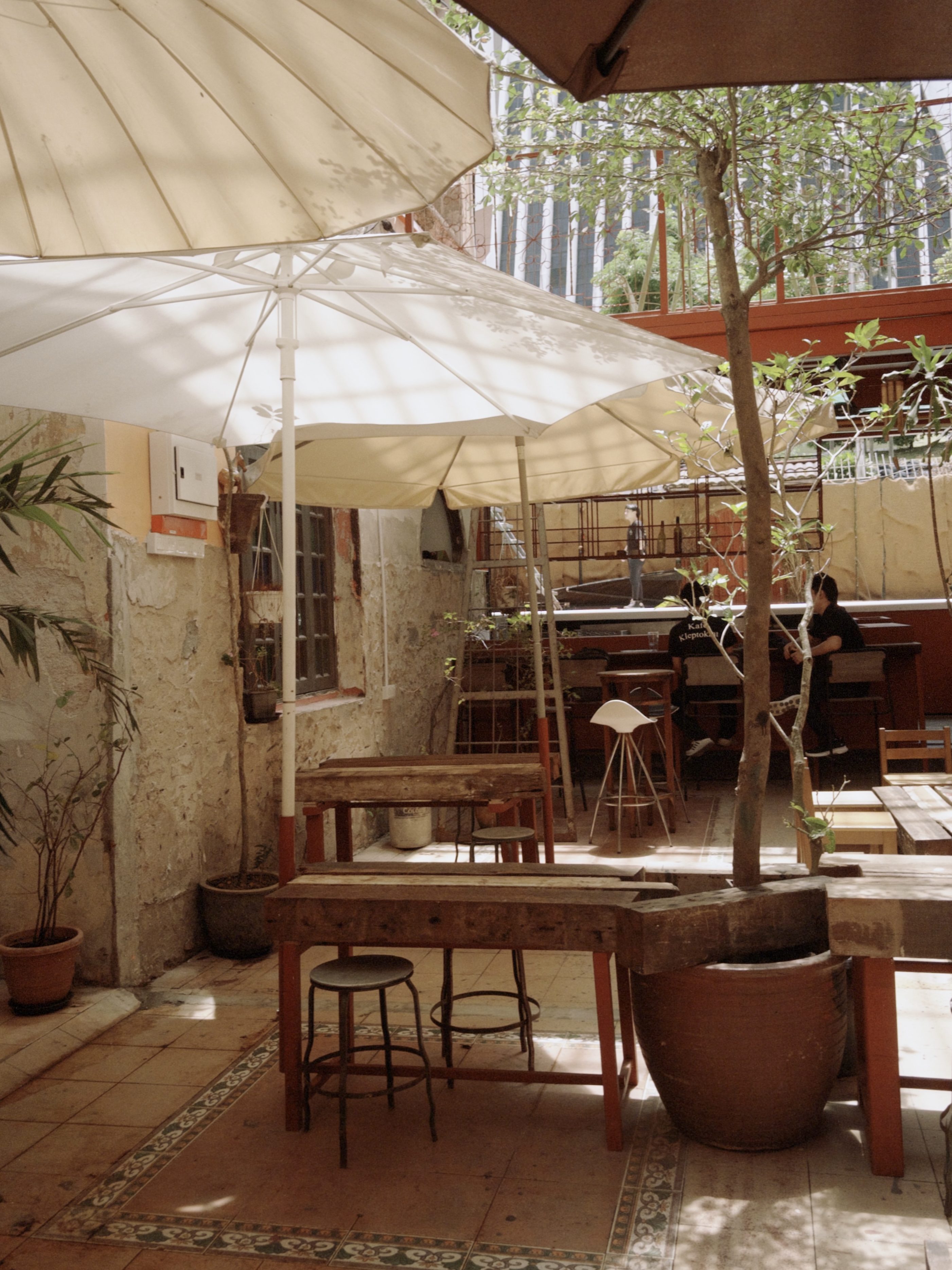 An inviting outdoor café space featuring wooden tables, mismatched chairs, and large umbrellas, surrounded by greenery and an old stone wall.