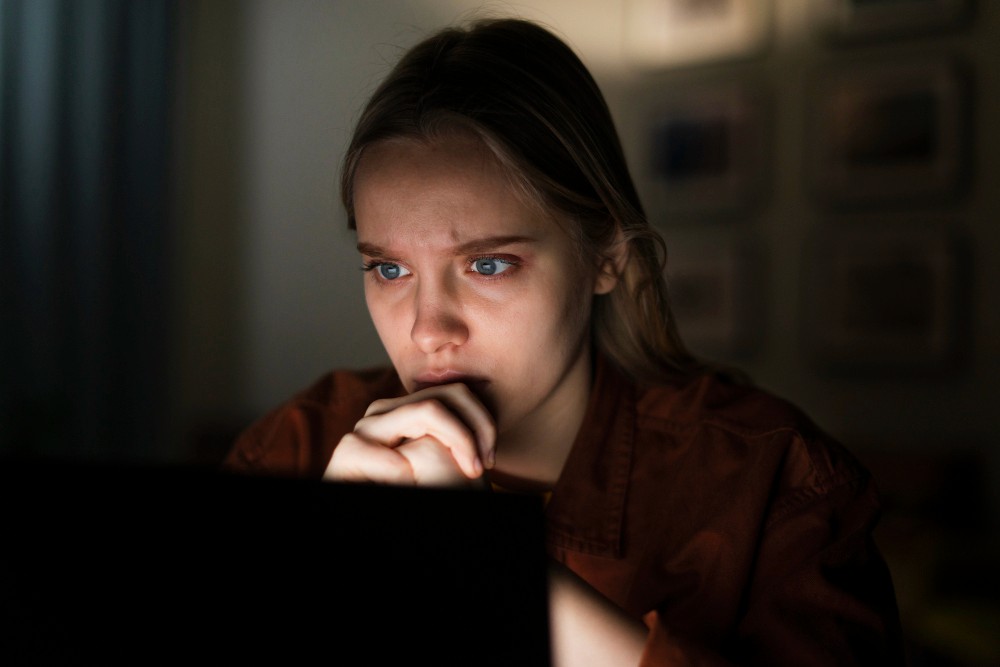 A young woman staring anxiously at a laptop screen in a dark room, illustrating digital anxiety and stress.