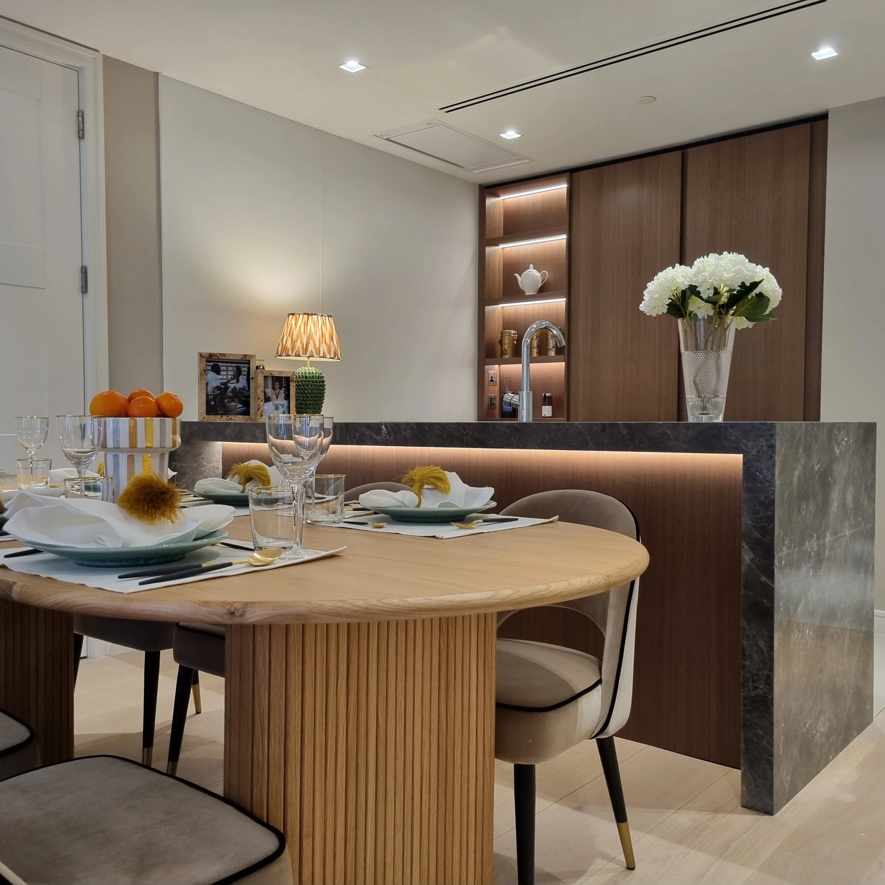 A modern dining area features a round wooden table with neatly set white plates, wine glasses, and a small bouquet of yellow flowers, complemented by a background of wooden cabinetry and a marble countertop displaying a vase of white blooms under warm ambient lighting.