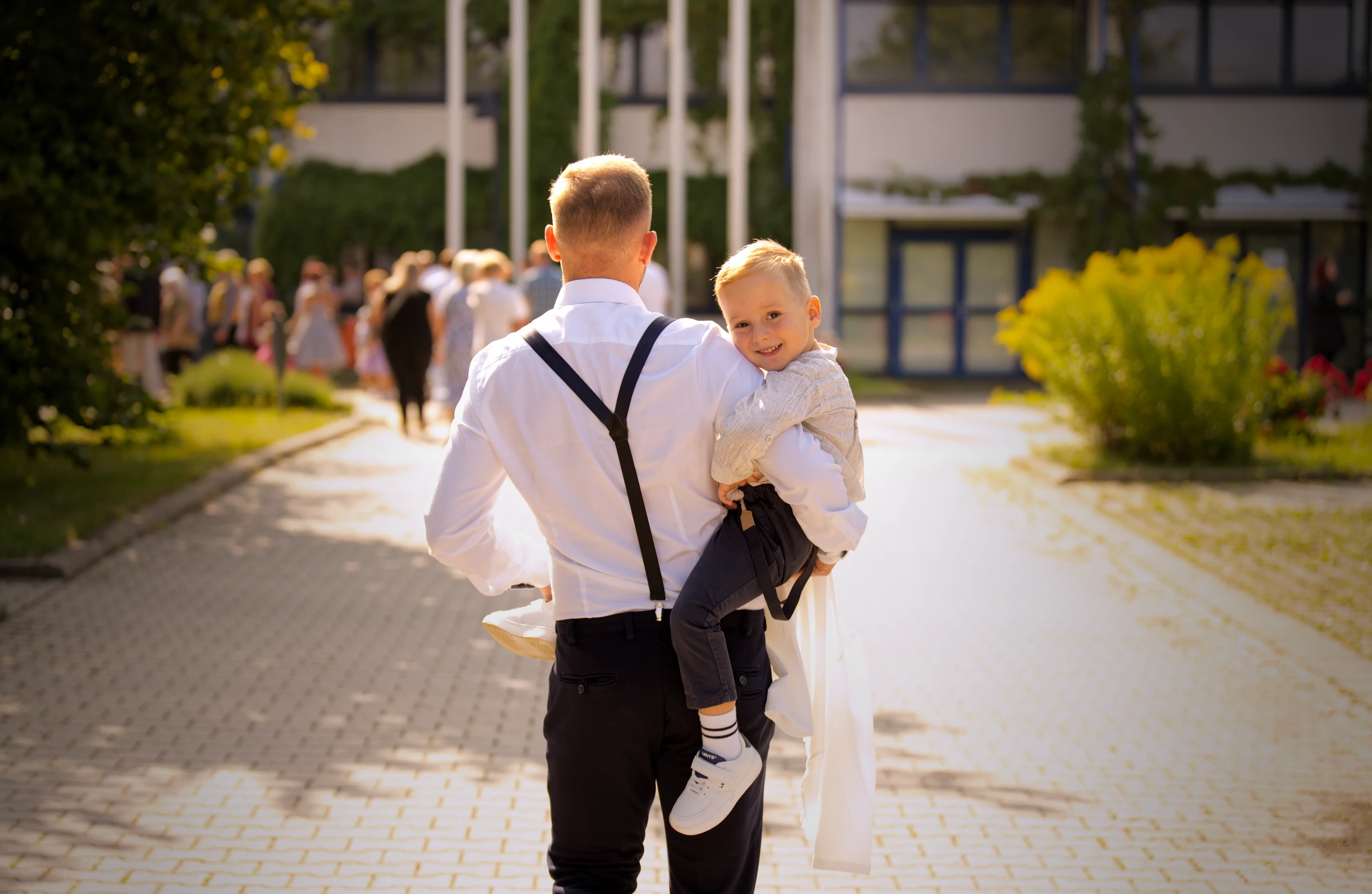 Emmys Vater trägt ihren kleineren Bruder über das Festgelände, während weitere Familien in das Gebäude der Einschulungsfeier gehen.