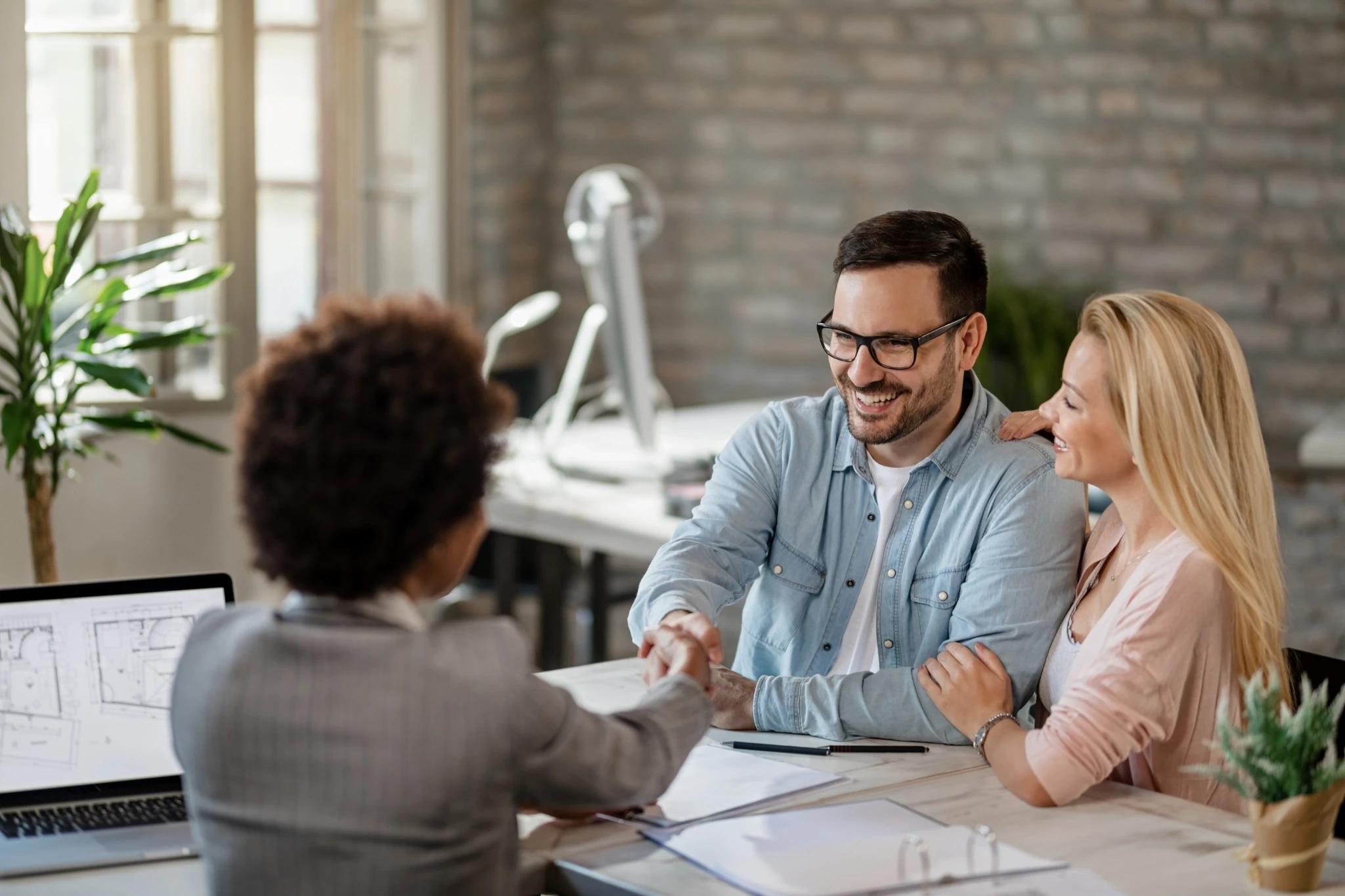 Three professionals engage in a handshake and friendly conversation at a modern office desk with laptops and architectural plans, surrounded by potted plants and brick walls with large windows.