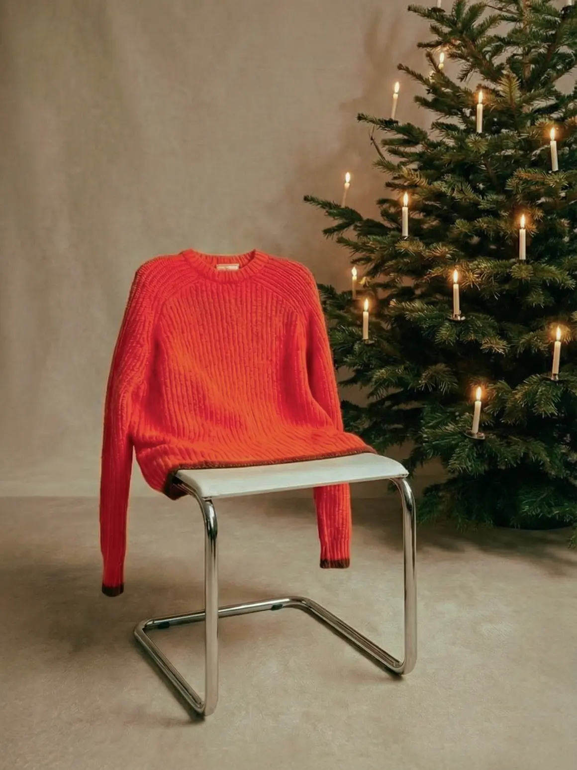 A vibrant red sweater is artfully draped over a sleek, modern chrome chair next to a festively adorned Christmas tree with lit candles, capturing a high-end visual for a fashion brand's AI production project.