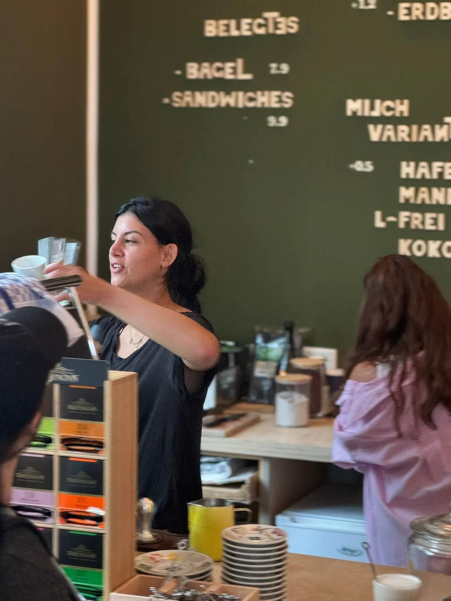 A woman stands behind a counter with various supplies, while another person is partially visible in the background.